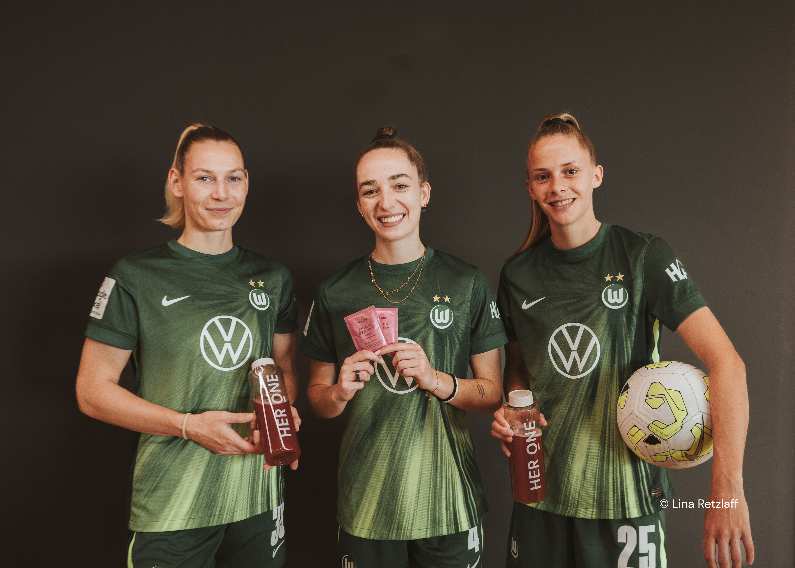 Trois joueuses du VfL Wolfsburg tiennent à bout de bras des bouteilles HERONELiquidBeauty et des sacs InnerBeautySachets.Football.