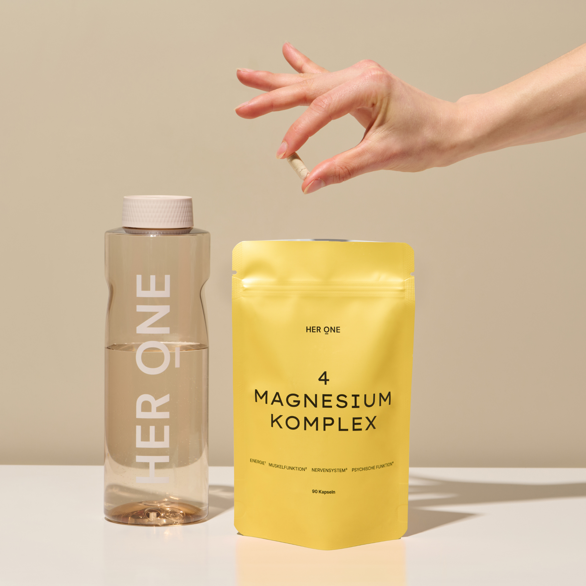 Une main tient une capsule au-dessus d'un emballage jaune de complexe de magnésium à côté d'une bouteille d'eau sur une table.