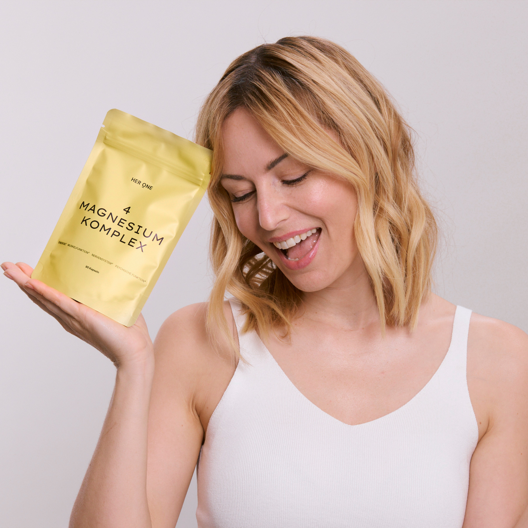 Une femme en haut blanc tient un sachet jaune HER ONE MAGNESIUM KOMPLEX et sourit.
