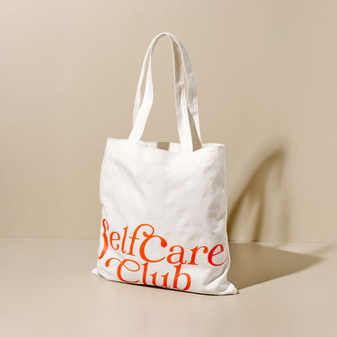 Borsa in tessuto bianco con scritta arancione "Self Care Club" su sfondo neutro.