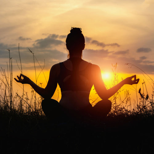 Persona in posizione di meditazione al tramonto, seduta su un sedile di loto in un prato e illuminata dal sole.