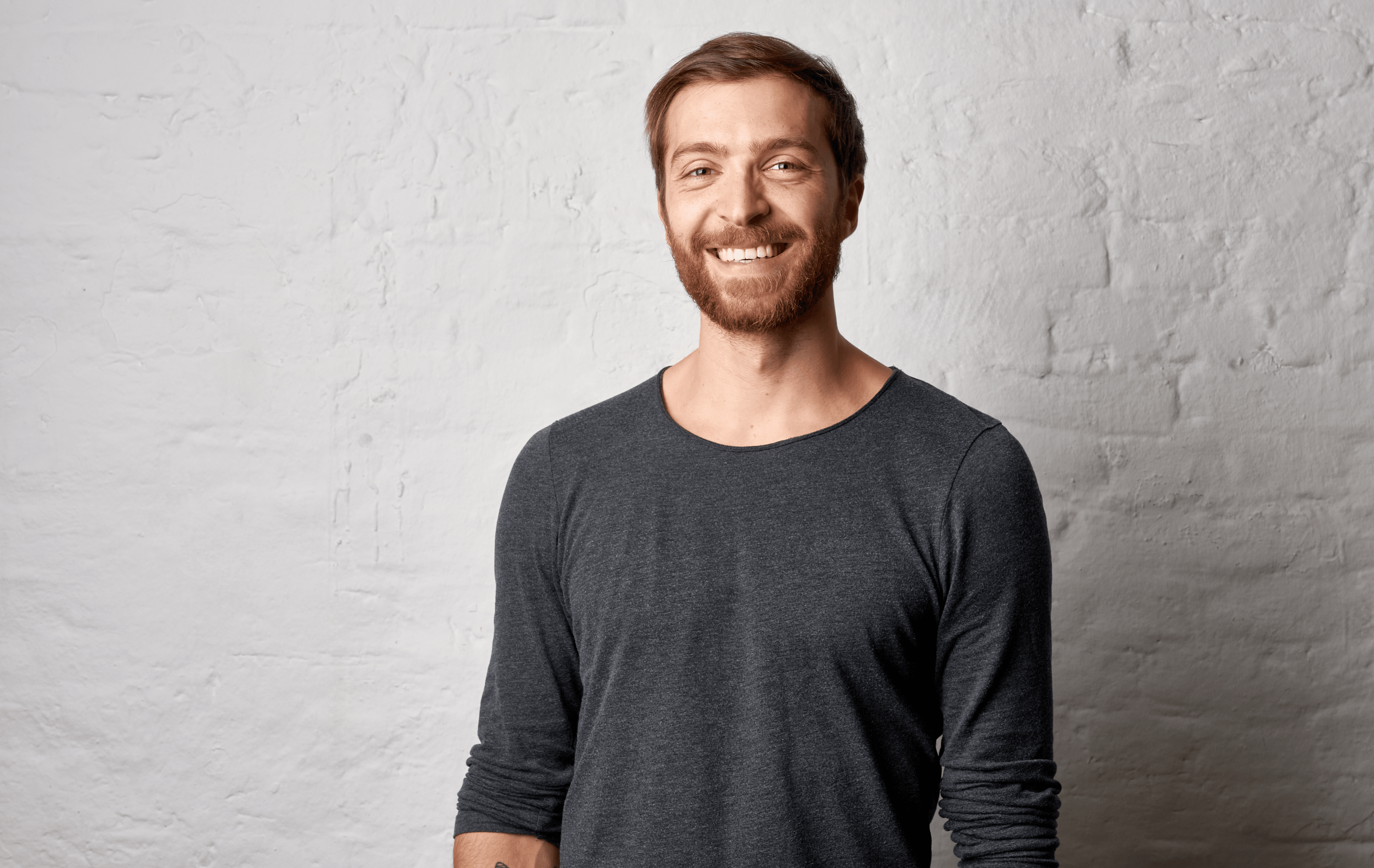 Uomo sorridente con la barba indossa una maglia grigia a maniche lunghe e sta in piedi davanti a un muro di mattoni bianchi.