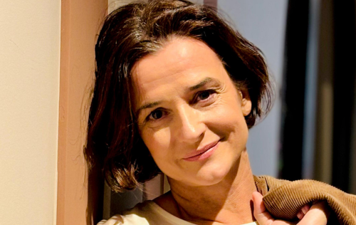 Una donna dai capelli castani si appoggia a un muro, sorridendo e tenendo in mano una giacca marrone.