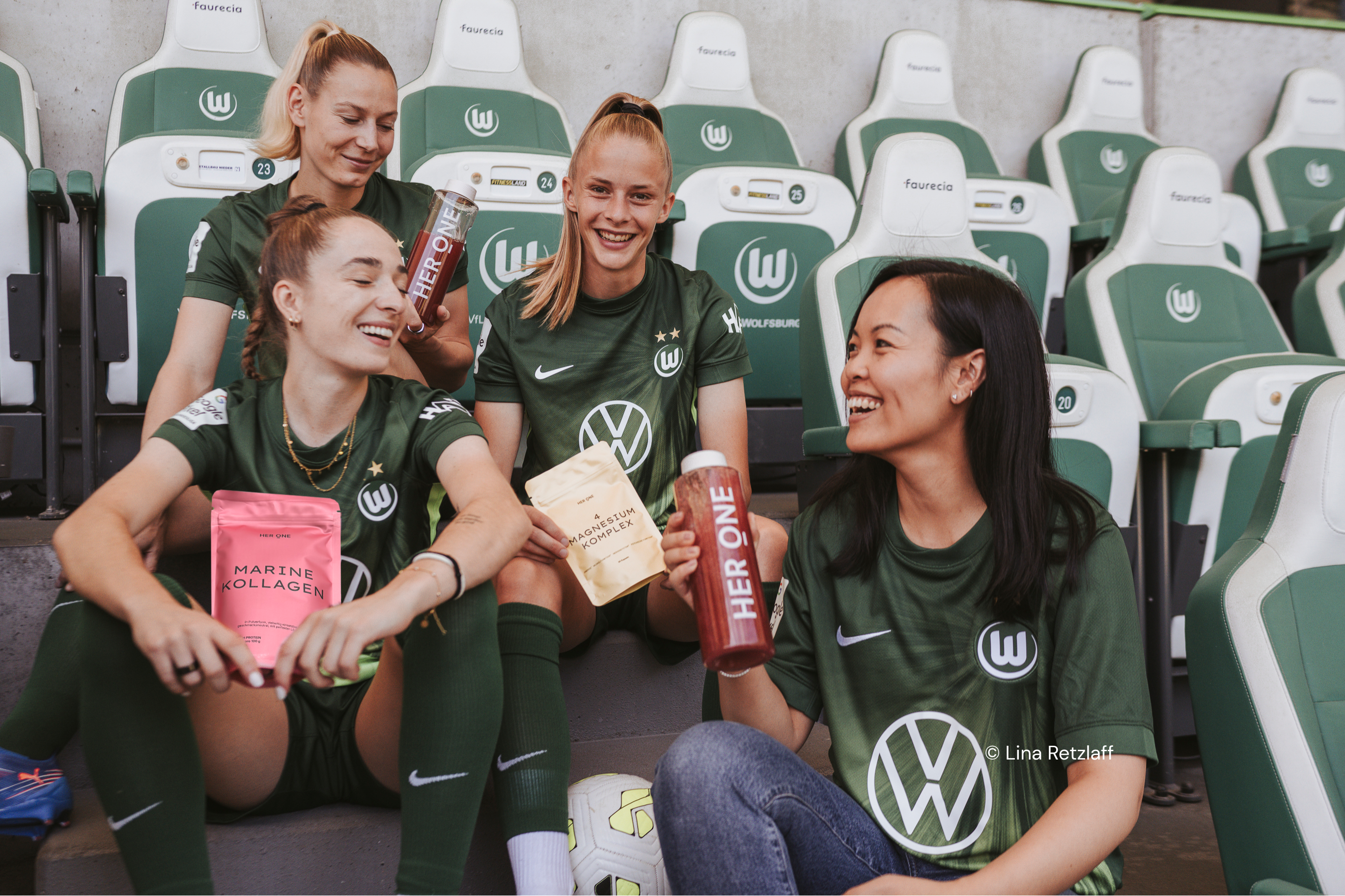Giocatori e donne del VfL Wolfsburg seduti sugli spalti, con in mano il complesso di collagene e magnesio HERONEMarine.