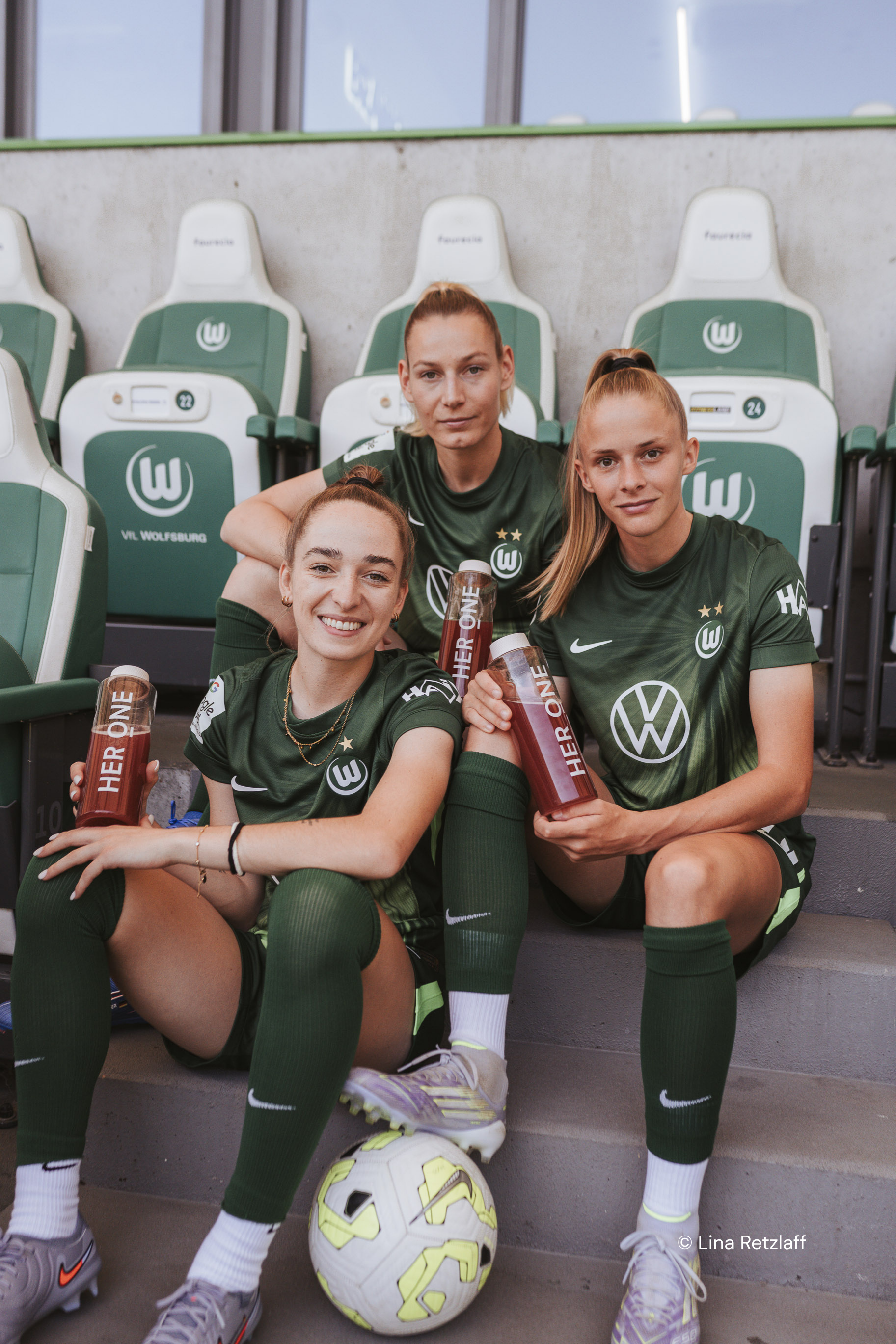 Tre giocatori del VfL Wolfsburg sono seduti sugli spalti, con in mano le bottiglie di bellezza HERONELiquid e indossando le maglie.