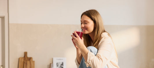 Vrouw met lang haar geniet van een glas rood sap terwijl ze glimlachend op een keukenblad zit.