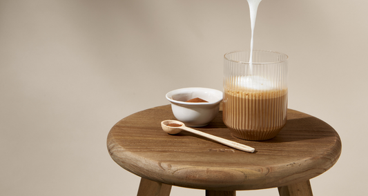 Een glas koffie met room wordt op een houten tafel gegoten, naast een lepel en een kommetje poeder.