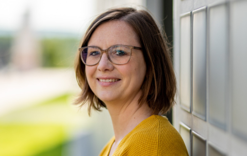 Een vrouw met een bril en een gele trui glimlacht en leunt tegen een muur.