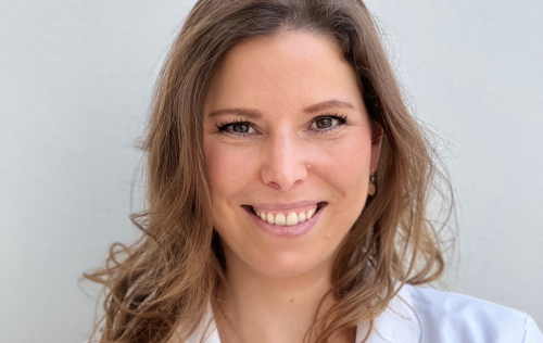 Een vrouw met lang bruin haar lacht naar de camera tegen een neutrale achtergrond.